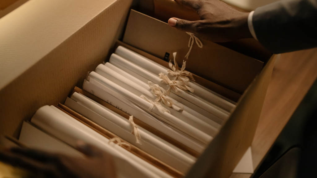 A person's hand reaches into a cardboard box filled with neatly organized documents tied with beige strings. The scene suggests careful organization and attention to detail.